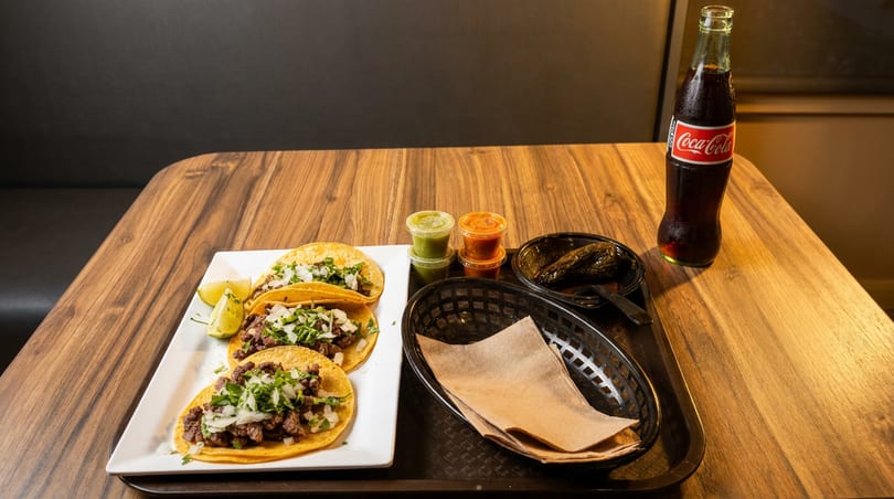 Three street tacos with lime, salsa, grilled peppers, and a Coca-Cola bottle on a wooden table.