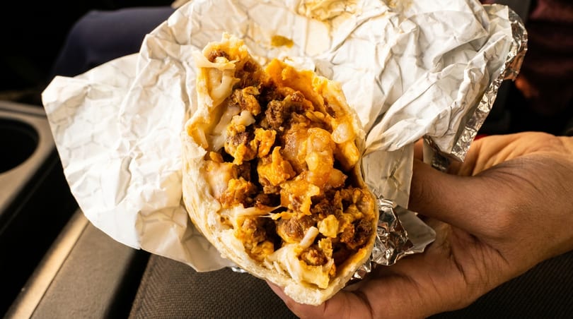A hand holds a large, open breakfast burrito filled with eggs, sausage, and cheese.