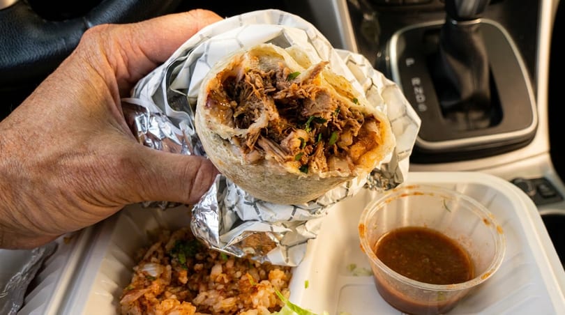 A hand holds a large, unwrapped burrito filled with meat and cilantro, with rice and salsa below.