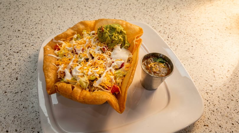 A taco salad in a crispy fried shell with guacamole, sour cream, and salsa on a white plate.