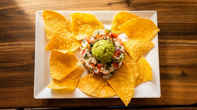 White plate with ceviche topped with guacamole, surrounded by tortilla chips on a wooden table.